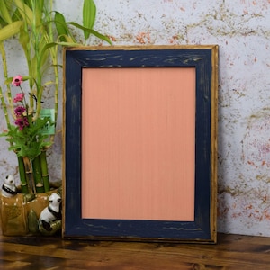 May include: A distressed wood picture frame with a dark blue inner border and a light peach colored mat. The frame is sitting on a wooden surface.