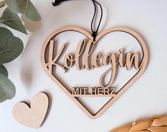 May include: A wooden heart-shaped ornament with the text "Kolleg*in mit Herz" in a cursive font. The ornament is hanging from a black string.