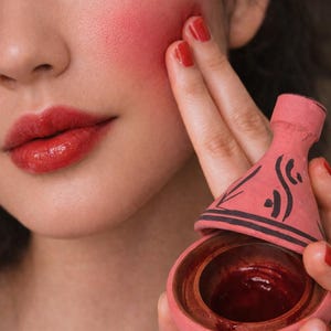Op de afbeelding: Gezicht van een persoon met rode lippenstift en blush. De persoon houdt een kleine, roze keramische container vast met een donkerrode substantie erin. De container heeft een decoratief deksel met zwarte accenten.