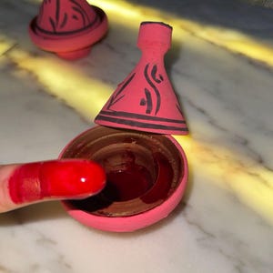 May include: Pink ceramic tagine pot with black decorative lines. The lid is off, revealing the interior. A finger covered in red paint is dipped into the pot. Another tagine pot is in the background.