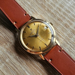 Può includere: Un orologio automatico Universal Polerouter Geneve color oro con cinturino in pelle marrone. Il quadrante dell'orologio ha una finitura dorata con lancette e indici neri. L'orologio è appoggiato su una superficie di legno.