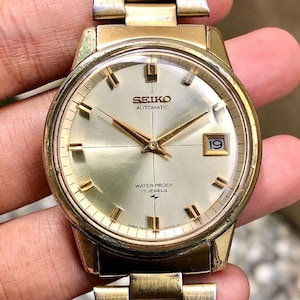 Op de afbeelding: Een vintage Seiko automatisch polshorloge met een goudkleurige kast en band. De wijzerplaat is licht champagnekleurig met gouden wijzers en markeringen. De datum wordt weergegeven op 3 uur. Het horloge is gemarkeerd met "WATER PROOF 17 JEWELS".