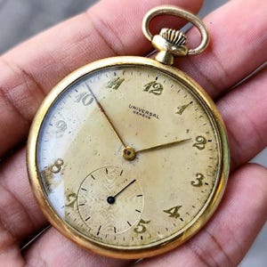 May include: A vintage gold-toned Universal Geneve pocket watch. The watch has a cream-colored face with black numerals and hands. A smaller dial is at the bottom. The watch is round with a loop at the top.