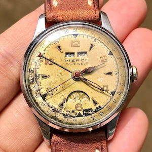 May include: A vintage Pierce watch with a gold-toned face, displaying the day and date. The watch has a brown leather strap and a silver-coloured case. The watch face shows the text "MON MAY" and "PIERCE 17 JEWELS".