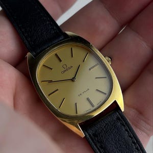 May include: A gold-toned Omega De Ville wristwatch with a black leather band. The watch face is rectangular with gold hands and markers. The watch has the Omega logo and "De Ville" printed on the face.