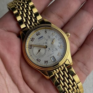 May include: A gold-tone Oris automatic wristwatch featuring a textured silver dial with Arabic numerals, a date window at the six o'clock position, and a multi-link bracelet. The watch face displays the text Oris Automatic and 27 Jewels.