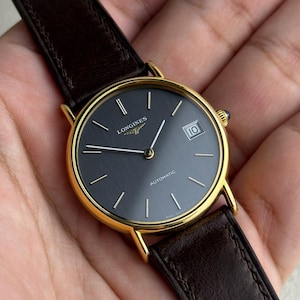 May include: A gold-toned wristwatch with a dark gray face and a brown leather band. The watch face displays the brand name "Longines" and the word "Automatic". The date is visible at the 3 o'clock position.