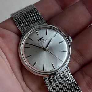 May include: A silver wristwatch with a mesh strap. The watch face has a brushed finish with black hour markers and hands. The brand name "IWC" is printed on the face. The watch is held in a hand.