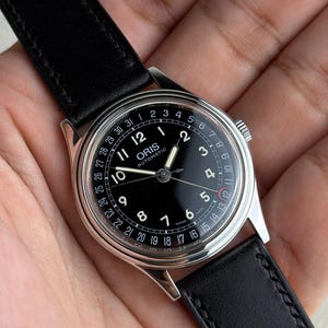 Könnte beinhalten: Eine Oris Automatic Armbanduhr mit schwarzem Zifferblatt, weißen arabischen Ziffern und einem Datumszeiger. Die Uhr verfügt über ein silberfarbenes Edelstahlgehäuse und ein schwarzes Lederarmband mit sichtbaren Nähten. Auf dem Zifferblatt steht Oris Automatic.