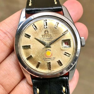 Puede incluir: Un reloj Titus Citomatic vintage con esfera dorada, correa de cuero negro y caja plateada. La esfera del reloj presenta marcadores de hora rectangulares, una ventana de fecha y el texto "Asian Games 1962 Souvenir Model."