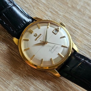 Puede incluir: Un reloj de pulsera automático Omega Genève Calendar dorado con esfera blanca y correa de cuero negro. La esfera muestra la fecha y el logotipo de Omega. Las manecillas y los marcadores de hora son dorados.