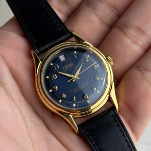 May include: A gold-toned wristwatch with a navy blue face and black leather band. The watch face features gold hands and hour markers, with the brand name "ORIS" visible. The watch has a date display at the 10 o'clock position.