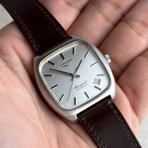 Könnte beinhalten: Eine silberne Longines Conquest Automatik-Uhr mit braunem Lederarmband. Die Uhr hat ein quadratisches Zifferblatt mit Datumsanzeige auf 3 Uhr. Das Zifferblatt ist silberfarben mit schwarzen Stundenmarkierungen und Zeigern.