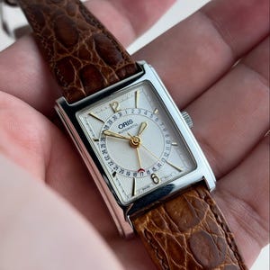 Może przedstawiać: Prostokątny srebrny zegarek na rękę z białą tarczą i brązowym skórzanym paskiem. Tarcza zegarka wyświetla datę i godzinę za pomocą złotych wskazówek i cyfr. Pasek ma fakturę przypominającą skórę krokodyla.