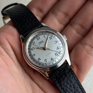 Może przedstawiać: Srebrny zegarek automatyczny Oris z jasną tarczą, czarnym skórzanym paskiem i wyświetlaczem daty. Tarcza zegarka posiada cyfry arabskie oraz napisy "ORIS AUTOMATIC" i "25 JEWELS".