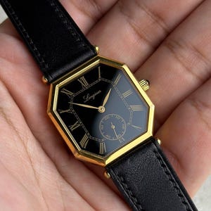 May include: A gold-tone octagonal wristwatch with a black dial, gold Roman numeral hour markers, and gold hands. The watch features the Longines logo in gold script on the dial, a small sub-dial for seconds, and a black leather strap with stitching.