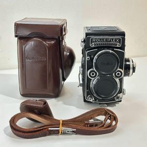 May include: A black Rolleiflex twin-lens reflex camera with two circular lenses, a brown leather hard case embossed with the Rolleiflex logo, and a separate brown leather camera strap with metal hardware and a small yellow rubber band.