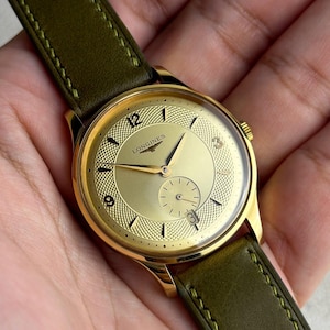 May include: A gold-tone Longines wristwatch featuring a textured gold dial with Arabic numerals at 12, 9, and 3 o'clock, a small seconds sub-dial, and a date window at 6 o'clock. The watch is attached to a dark olive green leather strap with stitching.