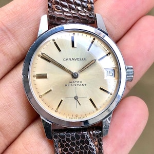Op de afbeelding: Een vintage Caravelle horloge met een zilveren kast en een goudkleurige wijzerplaat. Het horloge heeft zwarte uurmarkeringen, een datumvenster en de woorden "CARAVELLE" en "WATER RESISTANT". Het horloge heeft een bruine leren band.