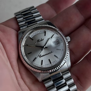 Peut inclure: Une montre Mido Commander argentée avec une lunette cannelée et un cadran argenté. Le cadran affiche le jour de la semaine, la date et le nom de la marque. La montre a un bracelet en acier inoxydable.