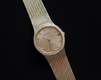 May include: A gold wristwatch with a woven gold band and a round face. The watch face is surrounded by a ring of small diamonds. The watch has a date display at the 3 o'clock position.