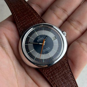 Könnte beinhalten: Eine Omega Genève Dynamic Automatik-Uhr mit silbernem Gehäuse, dunkelblauem Zifferblatt und braunem Lederarmband. Das Zifferblatt hat silberne Akzente und einen orangefarbenen Sekundenzeiger.