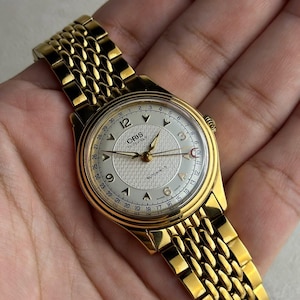 Puede incluir: Un reloj de pulsera dorado con una esfera texturizada y una pulsera dorada a juego. La esfera del reloj muestra la marca "ORIS" y la palabra "AUTOMATIC". El reloj tiene una visualización de la fecha alrededor del borde de la esfera.