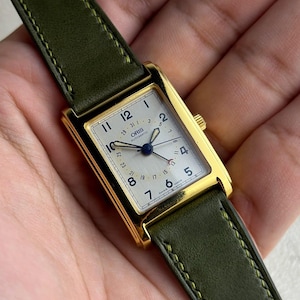 May include: A gold-toned rectangular wristwatch with a white face and black numerals. The watch has a green leather strap with yellow stitching. The watch face displays the brand "ORIS AUTOMATIC". The watch hands are blue.