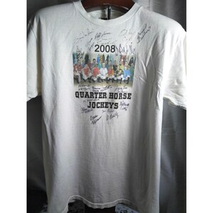 May include: Off-white t-shirt featuring a group photo of jockeys, the year "2008," and the text "Quarter Horse Jockeys." The shirt is adorned with multiple signatures. The jockeys are wearing colorful riding attire.