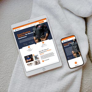 Op de afbeelding: Een witte tablet en smartphone die een website voor loodgietersdiensten weergeven. De tablet toont de volledige website-indeling, terwijl de telefoon een voor mobiel geoptimaliseerde versie weergeeft. De website toont de tekst: "Expert Plumbing Solutions for Homes & Businesses."