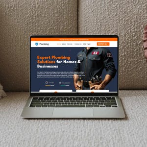 Op de afbeelding: Een zilveren laptop met een website voor een loodgietersbedrijf. De website bevat de tekst "Expert Plumbing Solutions for Homes & Businesses" en een afbeelding van een loodgieter. De laptop staat op een beige oppervlak.