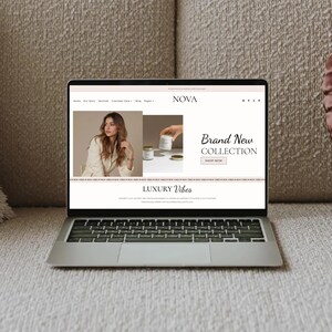 May include: A silver laptop displaying a website with the word "NOVA" at the top. The website features a woman in a cream-colored blazer, candles, and the text "Brand New Collection" and "Luxury Vibes". The laptop is on a beige couch.