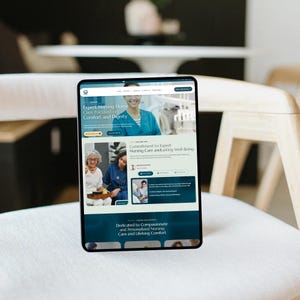 May include: A tablet displaying a website with the text "Expert Nursing Home Care Focused on Comfort and Dignity." The website features images of healthcare professionals and patients, with a teal and white color scheme. The tablet is resting on a white chair.