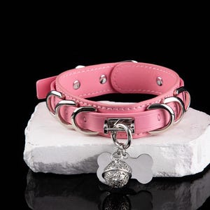 May include: Pink pet collar with silver-tone hardware. The collar features a decorative bell and a bone-shaped tag. The collar is made of a smooth material and has a buckle closure.