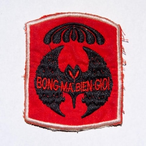 May include: A red and black embroidered patch with a bat-like emblem and text. The patch features a red background with a black bat design and the words "BONG MA BIEN GIOI" in red. The patch has a white border.