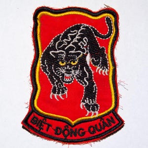 Può includere: Toppa ricamata con un disegno di pantera nera su sfondo rosso, con dettagli bianchi. La toppa ha un bordo giallo e nero e testo vietnamita. Questo è probabilmente un articolo militare o commemorativo.