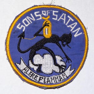 Pode incluir: Patch bordado circular com fundo azul e borda amarela. O patch apresenta as palavras "SONS OF SATAN" e "ALERE FLAMMAM". Uma figura negra semelhante a um demônio é retratada segurando um objeto amarelo.