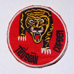 May include: Embroidered patch featuring a tiger with black and yellow stripes, set against a red background. The tiger is snarling, with white fangs visible. The patch has text in Vietnamese and English, and a white border.