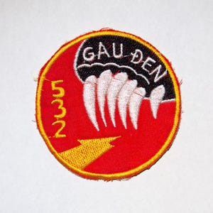 May include: A circular patch with a red background and a yellow border. The patch features the text "GAU DEN" in white, above a row of white claw-like shapes. The number "532" and a yellow arrow are also present.