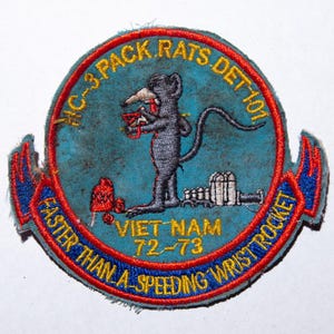 Peut inclure: Écusson brodé avec un fond bleu et une bordure rouge et jaune. L'écusson représente un rat de dessin animé tenant une fusée, avec le texte "MC-3 PACK RATS DET 101" et "Faster Than A Speeding Wrist Rocket".