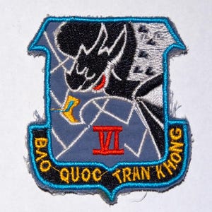 May include: Embroidered patch with a shield shape, featuring a black dragon-like figure, a red Roman numeral VI, and text in yellow and black. The patch has a blue border and a light blue background.