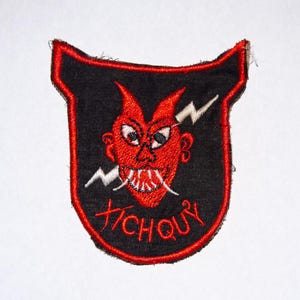 Könnte beinhalten: Gestickter Aufnäher mit schwarzem Hintergrund und rotem Teufelsgesicht mit weißen Fangzähnen und einem Blitz. Der Text "TICHQUY" ist rot am unteren Rand des Aufnähers gestickt. Der Aufnäher hat einen roten Rand.