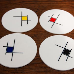 Può includere: Quattro sottobicchieri bianchi e rotondi con un design minimalista. Ogni sottobicchiere presenta un quadrato di colore diverso (giallo, rosso, blu e nero) con linee nere. I sottobicchieri sono disposti su una superficie di legno scuro.