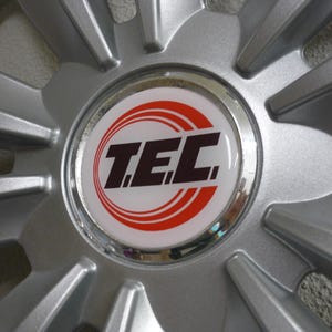 Op de afbeelding: Close-up van een zilveren autowiel met een centrale wieldop. De wieldop toont het woord "TEC" in zwarte letters, omcirkeld door twee rode ringen op een witte achtergrond. Het wiel heeft meerdere spaken die vanuit het midden uitstralen.