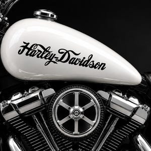 Op de afbeelding: Een witte motorfietsbrandstoftank met het zwarte Harley-Davidson-logo, geplaatst boven een gedetailleerde chromen motor met een ronde luchtfilterkap met een zesspaaks ontwerp en zwarte koelvinnen.