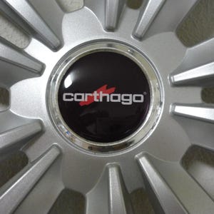 Könnte beinhalten: Silbernes Autorad mit radialem Speichendesign. Die Radkappe ist schwarz mit dem Wort "carthago" in Weiß und einem stilisierten roten Flammenlogo.