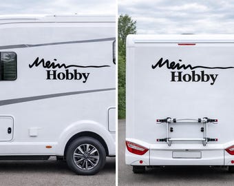May include: A white recreational vehicle featuring the black text Mein Hobby on the side and rear panels. The rear view displays a silver metal bicycle rack mounted on the exterior, along with red and white taillights and a license plate area.