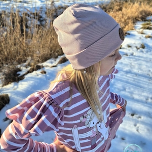 May include: A light mauve ribbed knit beanie with a small brown leather patch. The beanie is worn by a person wearing a pink and white striped long-sleeved shirt with a graphic print. The background shows snow and dry grass.