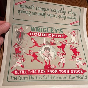 May include: A vintage Wrigley's Doublemint gum advertisement print on light gray paper. It features red-clad elf-like figures, a central portrait of a person, and text including 'Helps digestion, without question' and 'Refill this box from your stock'.