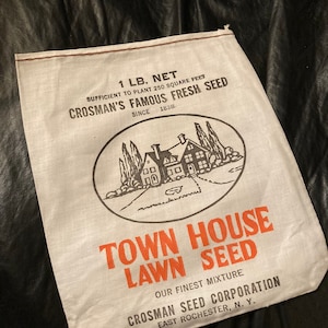 May include: A white burlap bag with red stitching, labeled "1 LB. NET" and "CROSMAN'S FAMOUS FRESH SEED" with a black and white illustration of a house and trees. The bag also reads "TOWN HOUSE LAWN SEED" and "OUR FINEST MIXTURE" in red text. The bottom of the bag reads "CROSMAN SEED CORPORATION EAST ROCHESTER, N. Y."